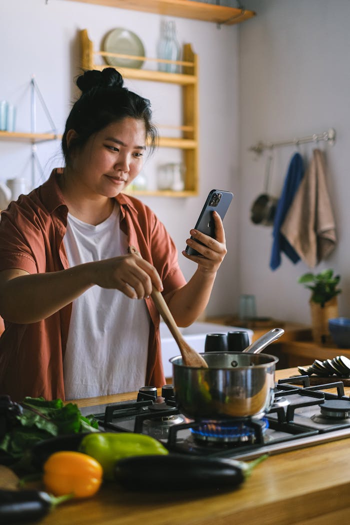 A woman uses her smartphone while cooking in a stylish kitchen, surrounded by fresh ingredients.