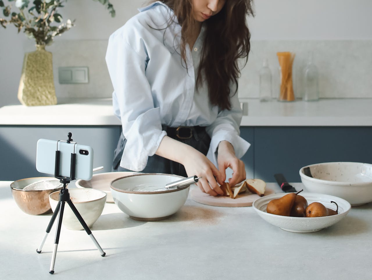 A woman preparing ingredients in a modern kitchen while recording with a smartphone on a tripod.