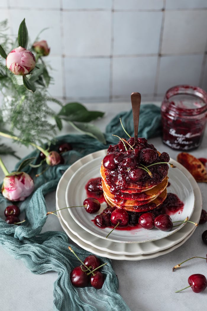Homemade pancakes topped with syrupy cherries, surrounded by flowers on a rustic table.
